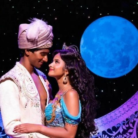 Two performers dressed as Aladdin and Jasmine stand close together on a stage, gazing at each other under a starry night backdrop with a large glowing blue moon and ornate palace-style set pieces behind them.