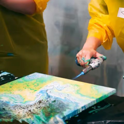A person wearing a yellow protective jacket using a small blowtorch to create fluid art on a canvas, with hands covered in paint and colorful abstract patterns forming on the artwork.