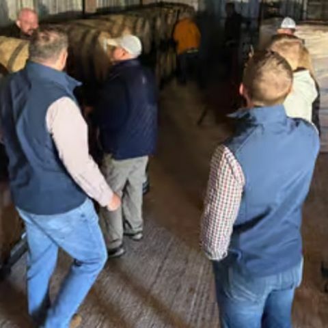 Group of people standing inside a distillery warehouse, gathered around stacked wooden bourbon barrels during a guided tasting tour.