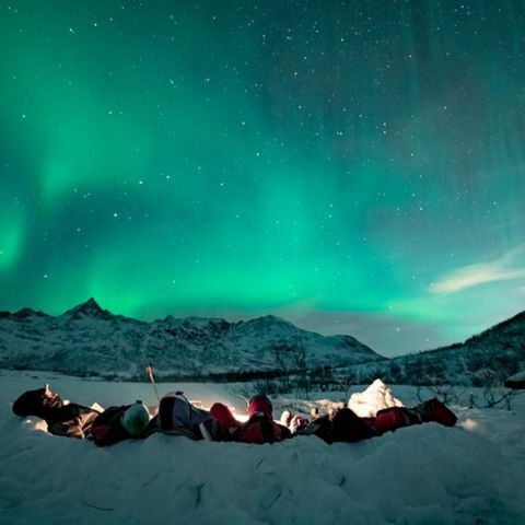 People lying on snow-covered ground, watching green Northern Lights glow across the night sky above snowy mountains.