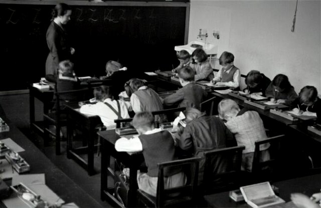 grayscale photography of teacher standing near chalkboard and children sitting on chairs