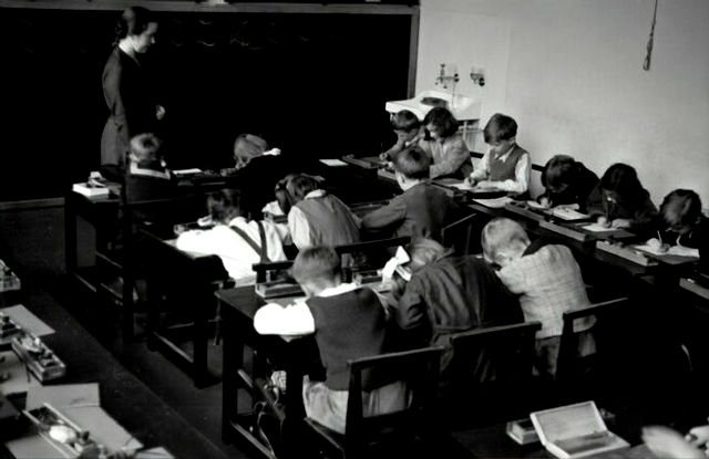 grayscale photography of teacher standing near chalkboard and children sitting on chairs