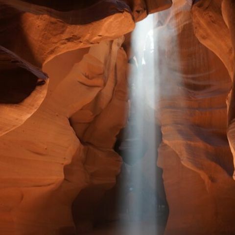 A narrow slot canyon with smooth, curved red rock walls, where a dramatic beam of sunlight pours down from an opening above, illuminating the canyon interior and creating a soft, glowing atmosphere.