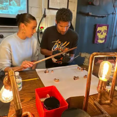 Two people working together at a workshop table, carefully measuring and assembling small components, surrounded by tools, copper fixtures, and glowing industrial-style lamps in a creative studio setting.