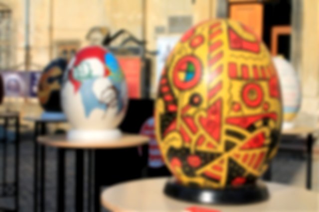 Large decorated eggs on stands, elevated on tables.  In focus one large yellow egg with modern orange and black patterns