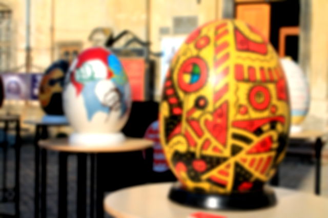 Large decorated eggs on stands, elevated on tables.  In focus one large yellow egg with modern orange and black patterns