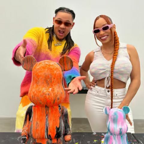 Two smiling artists standing behind paint-splattered bear-shaped art figures, posing playfully in a studio space, with one wearing a colorful sweater and the other dressed in white, both wearing sunglasses and presenting the finished artwork.