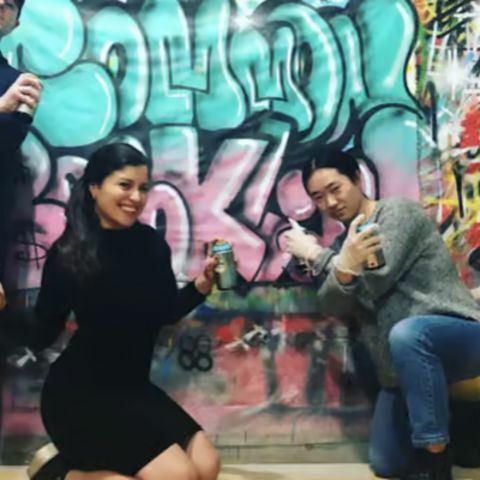 A group of friends posing playfully in front of a colorful graffiti-covered wall, holding spray paint cans and striking confident, fun poses in an urban street-art setting.