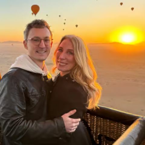 A couple smiling and embracing inside a hot air balloon basket at sunrise, with golden desert landscapes stretching below and several hot air balloons floating in the distance against a warm, glowing sky.