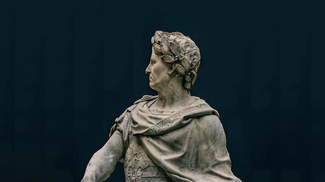 A close-up of the head and chest of the statue of Julius Caesar, facing left with a dark blue background
