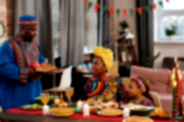 A celebratory meal, dressed in traditional african clothes a man stands holding a plate of food while a girl and woman sit at the table smiling