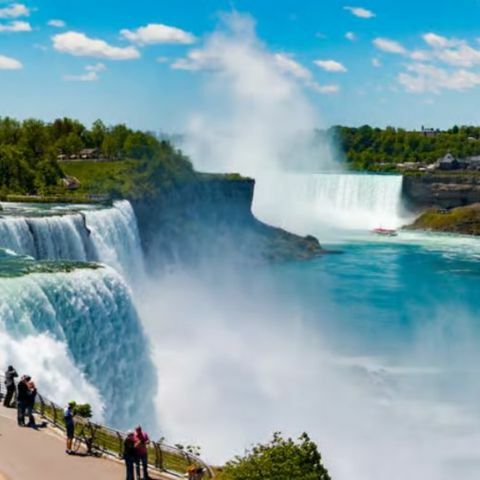 Niagara Falls with powerful waterfalls cascading into mist, visitors watching from a viewing path, and blue water under a clear sky.