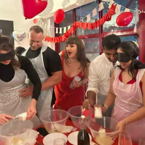 A lively group of people at a Valentine&rsquo;s-themed event, wearing aprons and blindfolds while mixing ingredients at a table, with red balloons, heart decorations, and one person in a red dress reacting excitedly in the background.