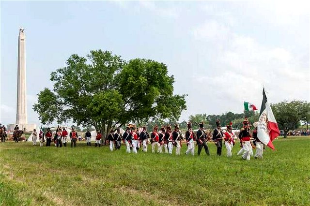 Soldiers marching reenacting a battle, passing through a green field in front of a tree.