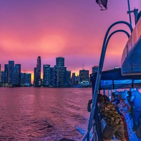 A city skyline at dusk viewed from a boat on the water, with tall buildings silhouetted against a vibrant pink and purple sky, rippling reflections on the surface, and passengers gathered along the boat&rsquo;s railing in the foreground.