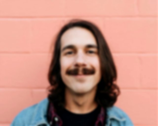 Man with moustache and long hair smiling in front of a pink wall