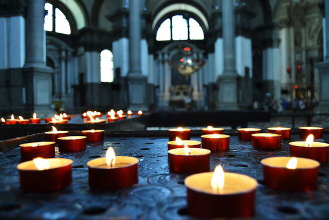 Multiple short red and gold candles stand on a table, soft blue blurry alter in the background