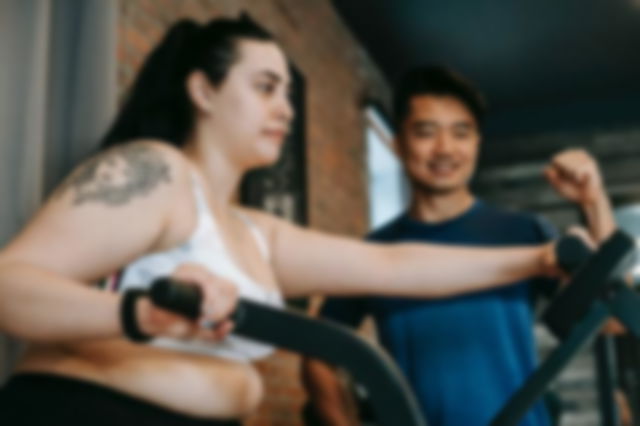 A large woman exercising, with tattooed arm, on a Gazelle machine while a smiling man supervises