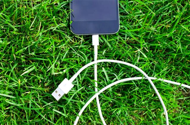 Iphone lying in bright green grass, turned off. Cable attached, but unplugged from the other side.