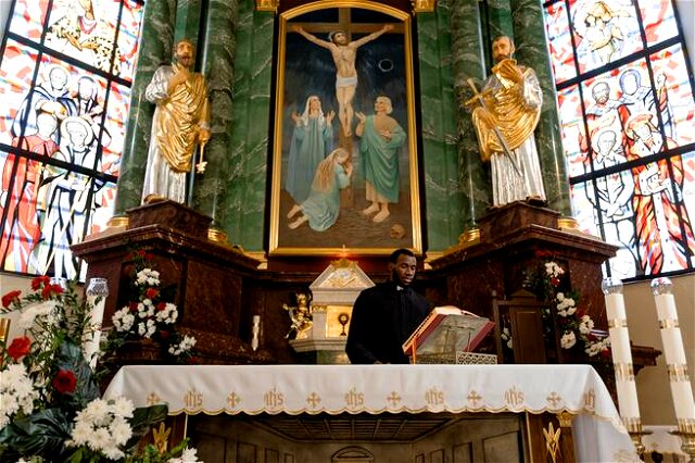 A priest standing at a pulpit, the cross featuring heavily in the centre of the shop, surrounded by colourful flowers and stained glass.
