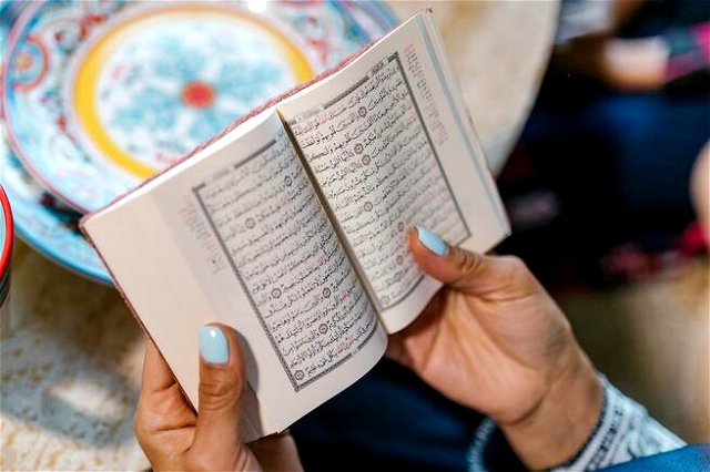 Two hands holding The Quran, thumb nails painted blue. Book text is decorative, colorful plate in the background.