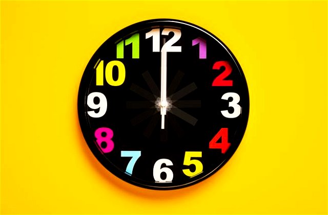 A black clock with colourful numbers lying in the middle of a bright yellow background.