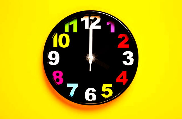 A black clock with colourful numbers lying in the middle of a bright yellow background.