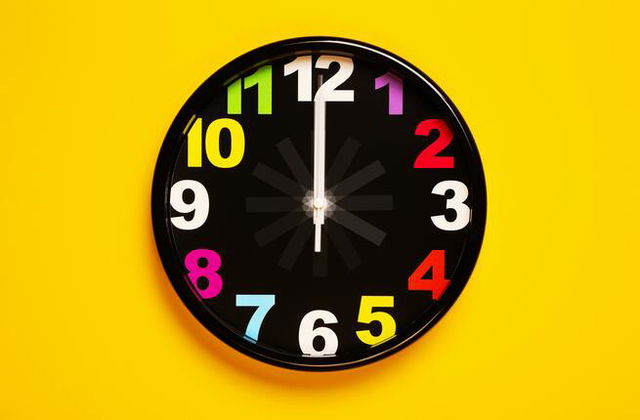 A black clock with colourful numbers lying in the middle of a bright yellow background.