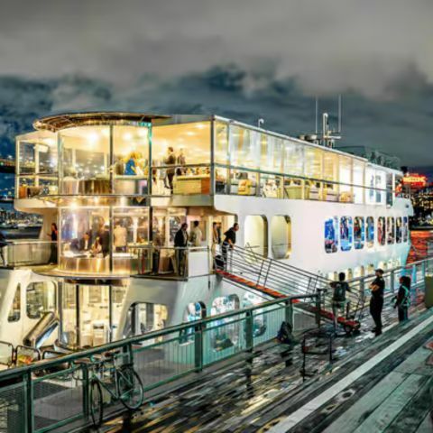 A large, brightly lit multi-deck cruise boat docked at a waterfront pier at night, with passengers visible through glass windows, people boarding via a ramp, city lights glowing in the background, and reflections shimmering on the water under a cloudy sky.