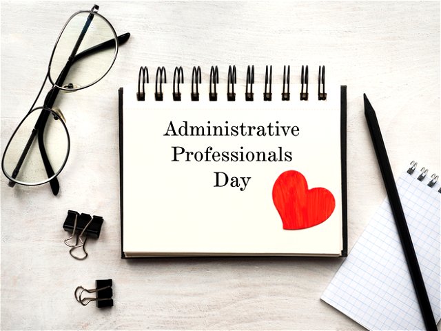 on a desk lies a clipboard that says ‘administrative professionals day’ with a heart, surrounded by clips, a pair of glasses and a pen