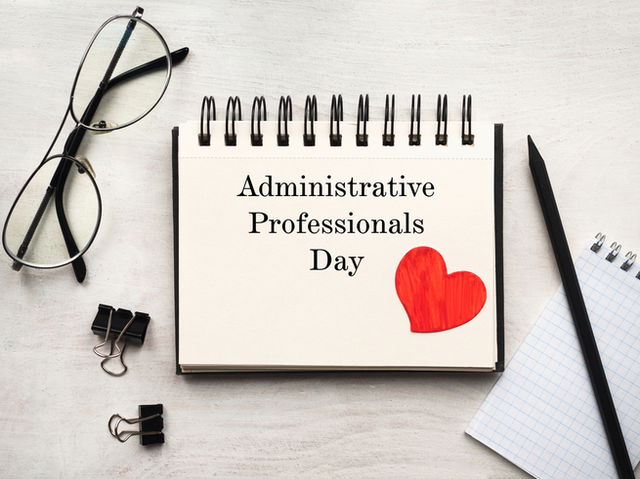 on a desk lies a clipboard that says ‘administrative professionals day’ with a heart, surrounded by clips, a pair of glasses and a pen