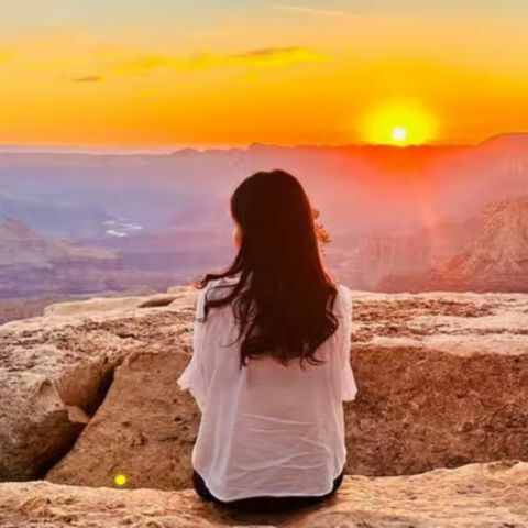 A woman sitting on a rocky cliff edge, facing away and looking out over a vast canyon landscape as the sun sets on the horizon, casting warm orange and golden light across the sky and layered cliffs below.