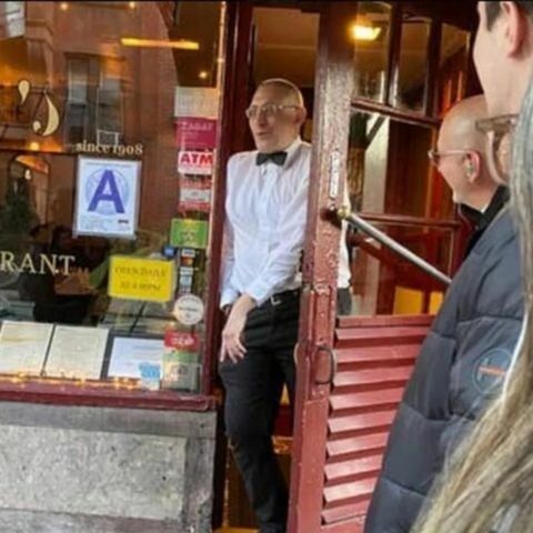 A waiter in a white shirt and black bow tie stands in the doorway of an Italian restaurant, speaking to people gathered outside, with menus displayed in the window and warm indoor lighting visible behind him.