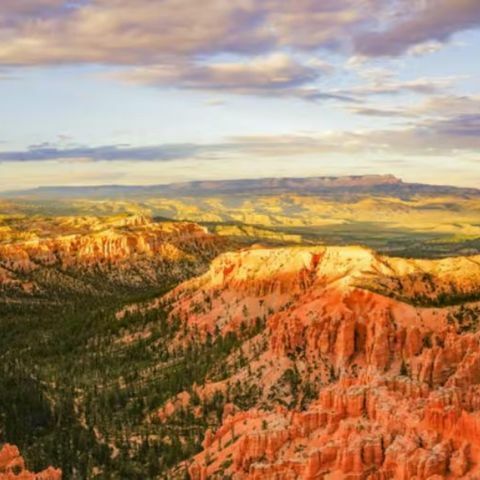 Wide view of Bryce Canyon with red rock hoodoos, forested valleys, and layered cliffs glowing under a colorful sky.