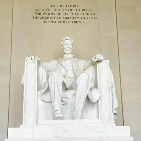The white marble statue of Abraham Lincoln seated in the Lincoln Memorial, with his hands resting on the chair arms and an inscription carved high on the wall behind him.