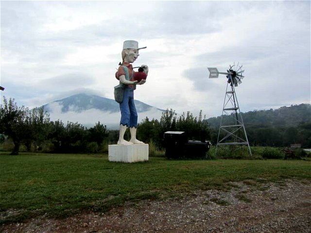 The statue of Johnny Appleseed in front of a weathervane on a dull day, clouds and hills in the background