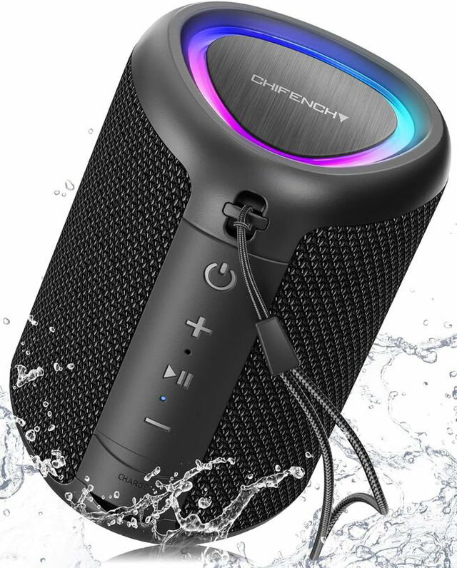 A black portable Bluetooth speaker with textured fabric sides, illuminated LED ring on top, control buttons on the front, and a wrist strap attached, shown splashing through water to highlight its waterproof design.