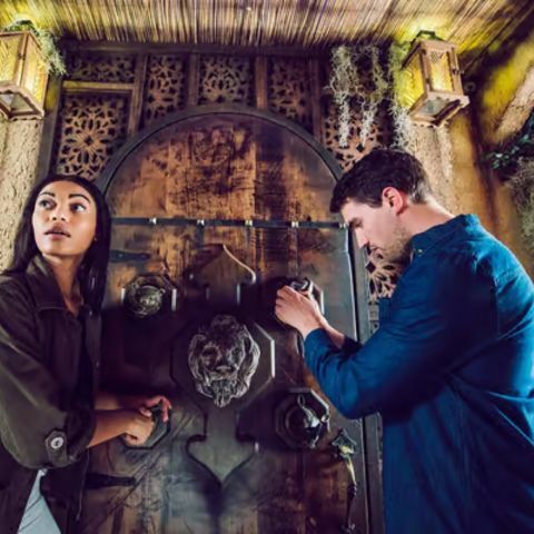 Two people standing inside an immersive escape-room setting, examining a large ornate wooden door with carved details and locks, lit by warm lanterns, as one person looks around alertly while the other works to solve a puzzle.