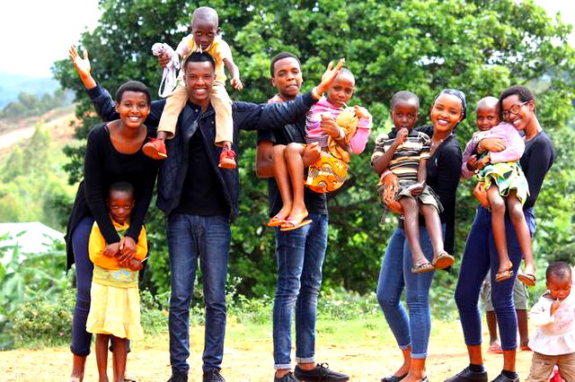 Image of large family, five adults, four of which are carrying children. Standing outside on sandy path with a tree in the background