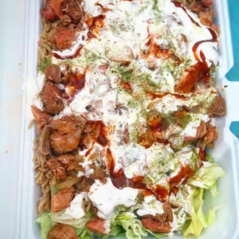 An overhead view of a takeout container filled with a mixed rice and salad dish topped with grilled meat, drizzled with creamy white sauce and reddish seasoning, set against a bright blue background.