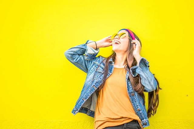 A young lady with a large smile against a yellow background. She is wearing a denim jacket and orange teeshirt.