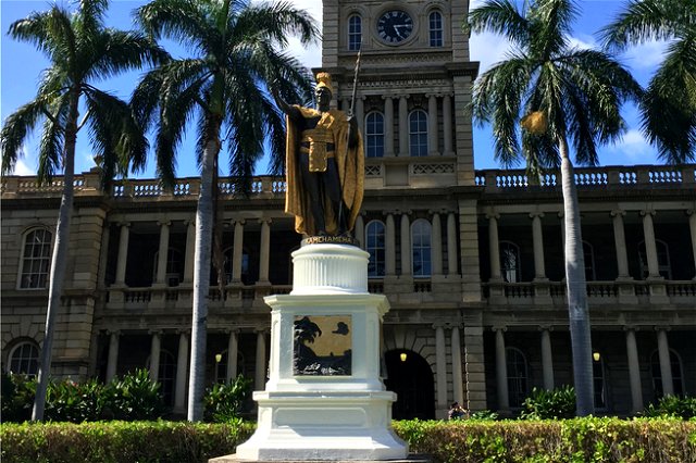 a golden statue on a plinth, with bushes and palm trees and a building behind it