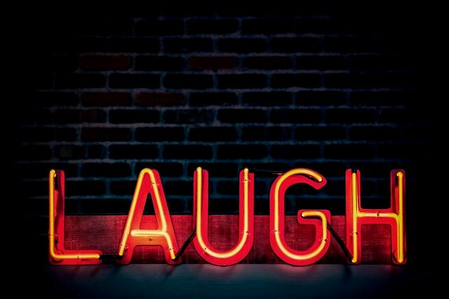 A red neon sign that reads ‘laugh’ against a dark brick wall. The corners of the image with dark vignetting.