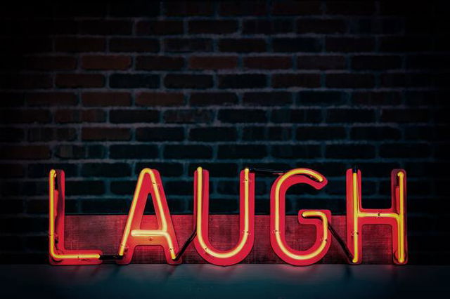 A red neon sign that reads ‘laugh’ against a dark brick wall. The corners of the image with dark vignetting.