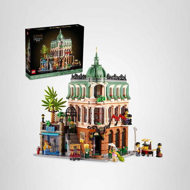 A detailed LEGO Icons Boutique Hotel building set with a pastel green dome and ornate architecture, shown assembled with minifigures, street details, and accessories, alongside the product box in the background.