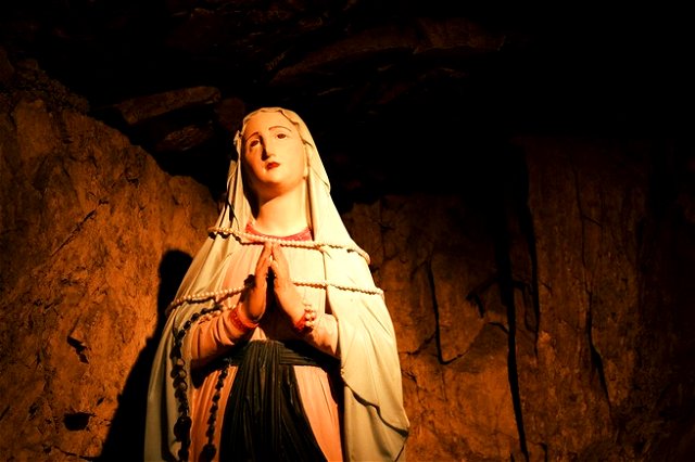 A statue of mother mary, hands together praying stones of a cave behind her.