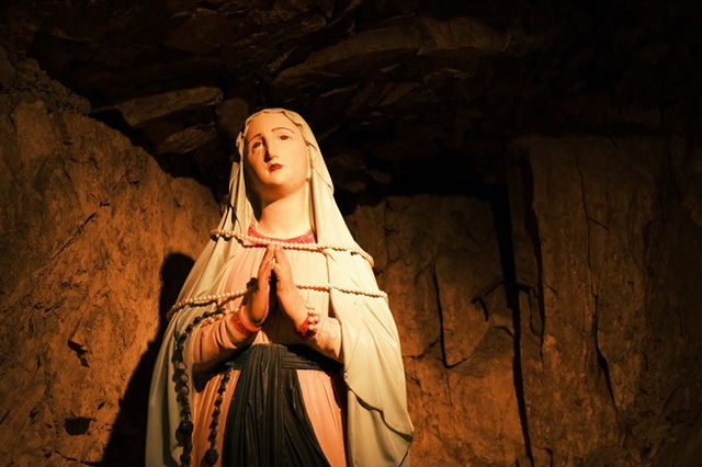 A statue of mother mary, hands together praying stones of a cave behind her.