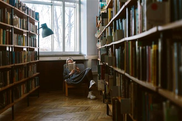In an ancient library a woman is sat reading at the end of a corridor of books on shelves.