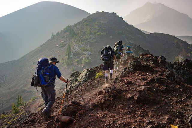 A group of hikers walk along the edge of a hill in a dark brown, arid, dusty landscape