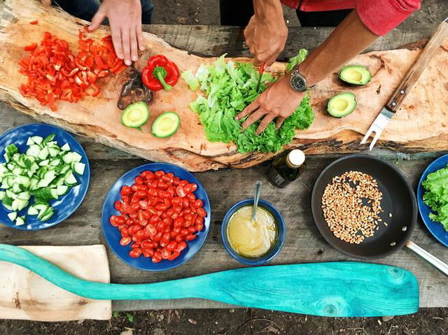 Two people at a wooden chopping board, preparing vegetable and salads in blue bowls